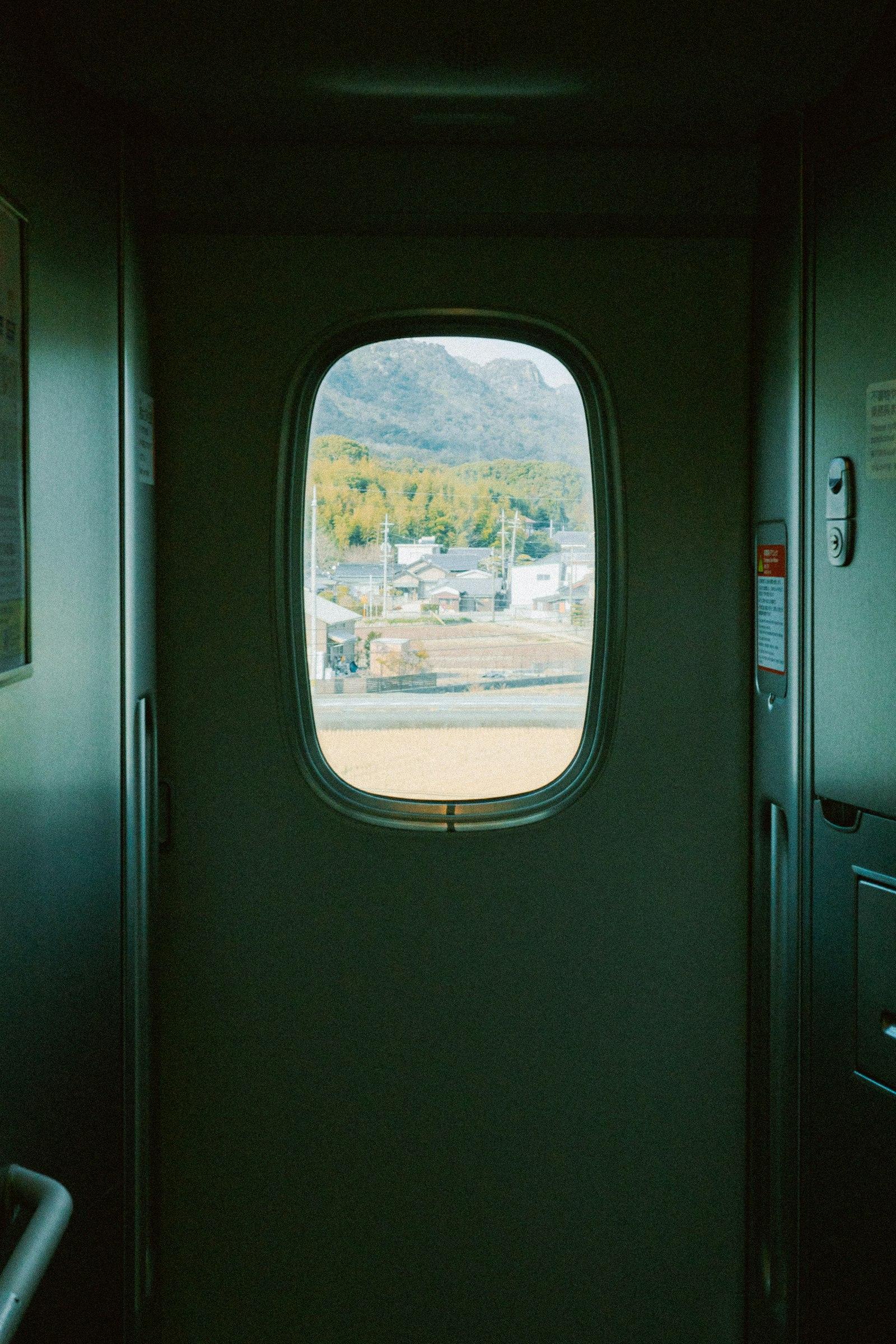 A porthole-shaped train window looking out over Japanese countryside from a dim train corridor